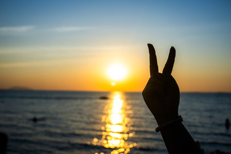 silhouetted hand making a peace sign against a glowing sunset over the ocean, with golden light reflecting on the waterの写真素材