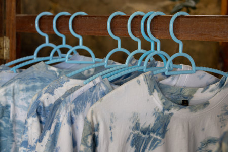 close-up shot of tie-dye t-shirts hanging on light blue plastic hangers. The shirts feature a soft, pastel tie-dye pattern. The hangers are neatly arranged on a wooden rackの写真素材