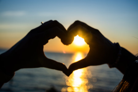 Silhouetted hands forming a heart shape against a golden sunset over the ocean, with sunlight reflecting on the calm water.の写真素材