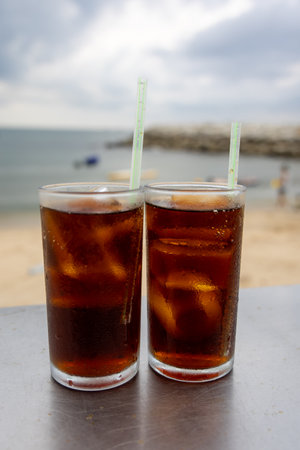 Two glasses of iced cola sit on a table by the sea, with a cloudy sky backdrop. The drink's bubbles...