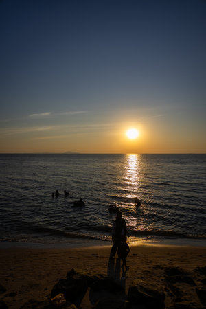 breathtaking sunset over a calm ocean, with the sun casting golden reflections on the water and silhouettes of people enjoying the tranquil scene near the shorelineの写真素材