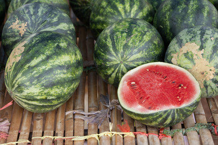 several fresh watermelons displayed on a bamboo mat at a market. One watermelon is cut open, revealing its juicy red flesh and black seeds. The image captures the vibrant colors and freshness of the fruit.の写真素材