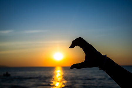silhouetted hand forming a half-heart gesture in front of a glowing sunset over the ocean, with golden sunlight reflectingの写真素材