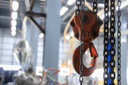 A close-up of a red industrial chain hoist with heavy-duty metal chains, suspended in a factory or workshop environment. The blurred background showcases machinery and equipmentの写真素材