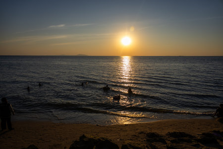 breathtaking sunset over a calm ocean, with the sun casting golden reflections on the water and silhouettes of people enjoying the tranquil scene near the shorelineの写真素材