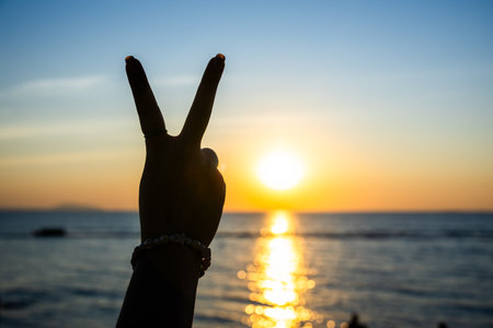 silhouetted hand making a peace sign against a glowing sunset over the ocean, with golden light...