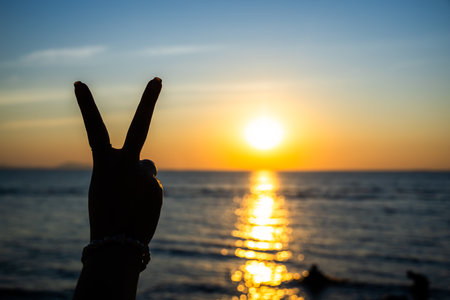silhouetted hand making a peace sign against a glowing sunset over the ocean, with golden light reflecting on the waterの写真素材