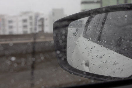 close-up of a car's side mirror covered in raindrops, reflecting a blurred urban scene with buildings in the background.の写真素材