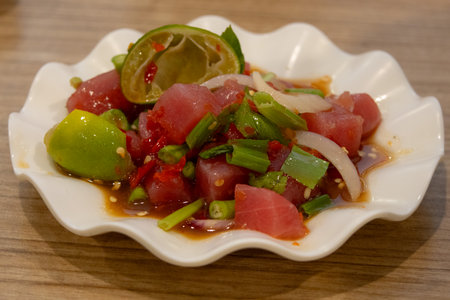 Close-up of a white plate with diced raw tuna, chili peppers, green onions, sliced onions, and lime wedges in a savory sauceの写真素材