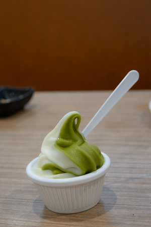 small cup of swirling matcha and vanilla soft serve ice cream, with a white plastic spoon on a wooden table.の写真素材