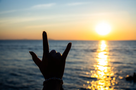 silhouetted hand making the "I Love You" sign in front of a radiant sunset over the ocean, with golden sunlight reflecting on the water.の写真素材