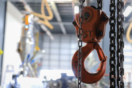 A close-up of a red industrial chain hoist with heavy-duty metal chains, suspended in a factory or workshop environment. The blurred background showcases machinery and equipmentの写真素材
