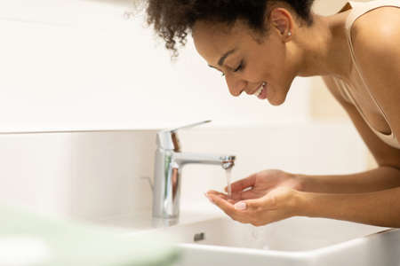 African American woman washes her face with water at the bathroom sink.