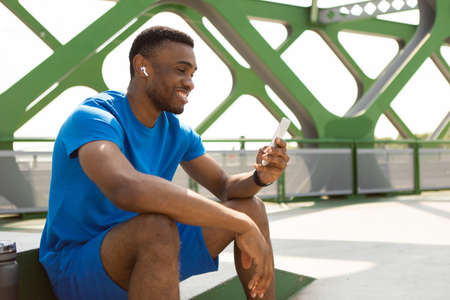 Cheerful african american sportsman with wireless headphones using smartphone while relaxing on park...