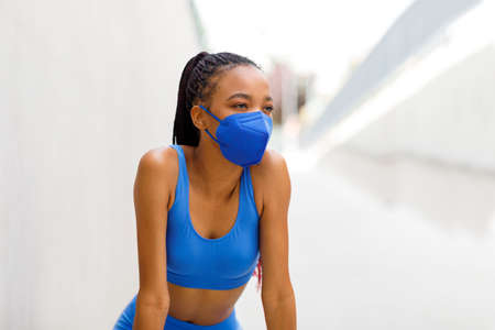 A black woman runner stands in a mask from the coronavirus, bending over and taking a breath after a runの写真素材