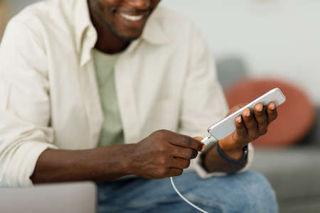 A young man sits on a sofa and charges a mobile phone battery using a wire.の写真素材