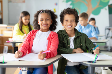 Happy black schoolboy and schoolgirl looking at camera and smiling, sitting at desks with other children on backgroundの写真素材