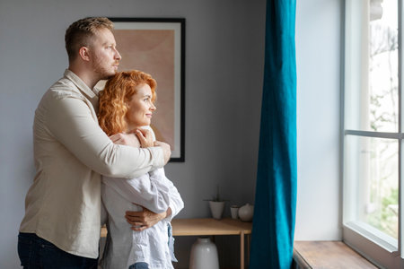 Young couple in love hugging standing near window at home, cuddling and enjoying time together, side view, free spaceの写真素材