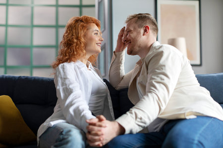 Young romantic European couple enjoying talk at home, sitting on sofa, holding hands and smiling at each otherの写真素材