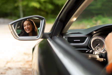 Beautiful young European woman sitting in her new car and looking throw the back view mirror and...