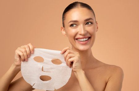 Beautiful happy European lady using moisturizing sheet face mask, posing isolated on beige background. Skin care, cosmetology, beauty conceptの写真素材