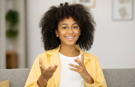 Portrait of young happy African American woman sitting on sofa at home and smiling at camera, gesturing with her hands during video callの写真素材