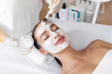 Beauty treatment. Professional beautician applying clay mask to lady at spa salon, woman enjoying face care procedures, above viewの写真素材