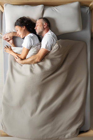 Bedtime. Spouses sleeping, lying on big bed and resting, husband embracing wife from back, top view,...