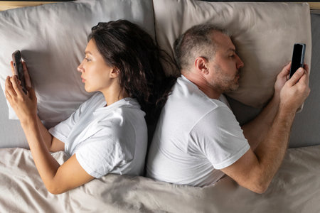 Indifference in relationships. Spouses using cellphones lying back-to-back in bed, ignoring one...