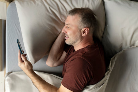 Positive European middle aged man scrolling in bed, using cellphone, holding gadget and smiling, top...