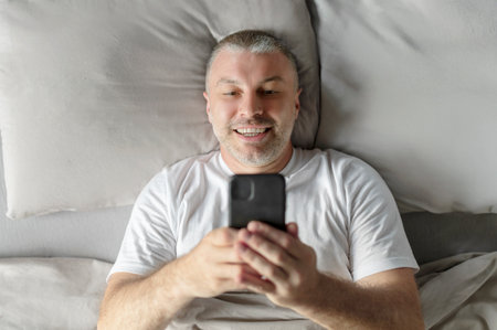 Happy middle aged man laying in bed after waking up in the morning and checking cellphone, chatting with lover and smiling, top viewの写真素材