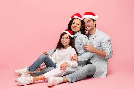 Adorable happy European parents and their daughter posing in red Santa hats over pink studio...