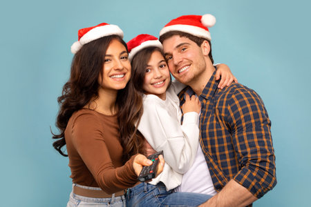 Portrait of affectionate European parents embracing with daughter, wearing Santa hats hugging and smiling at camera over blue backgroundの写真素材