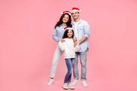 European family in Santa hats, hugging and smiling at camera, posing against pink background, full length shot. Celebrating winter holidays togetherの写真素材