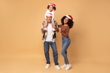 Happy young black parents and little daughter sitting on father shoulders, family in Santa hats posing on beige background, full lengthの写真素材