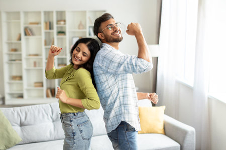 Joyful indian couple dancing in living room interior, having fun and singing, husband and wife enjoying spending time togetherの写真素材
