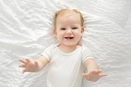 Cute European baby girl in white bodysuit lying on bed on her back and smiling at camera, top view shotの写真素材