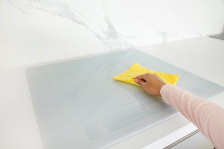 Female hand cleaning modern white induction hob by yellow rag, doing housework on weekend, free...