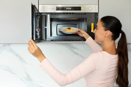 Woman heating spaghetti in microwave oven, using modern kitchen appliance for cooking food, preparing lunch or dinnerの写真素材