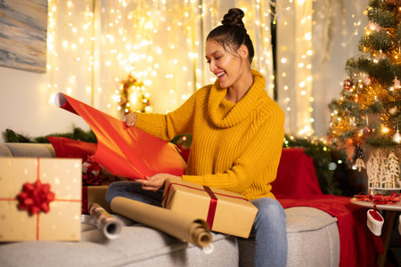 Positive young woman getting ready for Christmas and wrapping gift boxes, using red paper for wrapping, sitting on sofa in decorated roomの写真素材