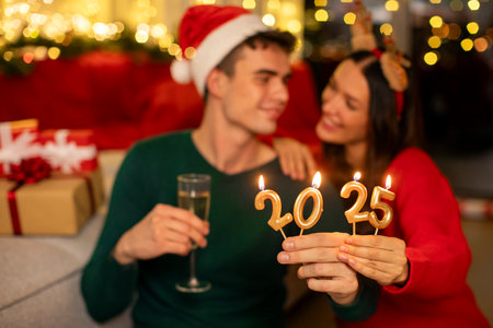 Focus on 2025 number candles in hands of young couple celebrating New Year and holding glasses of champagne in Xmas decorated roomの写真素材