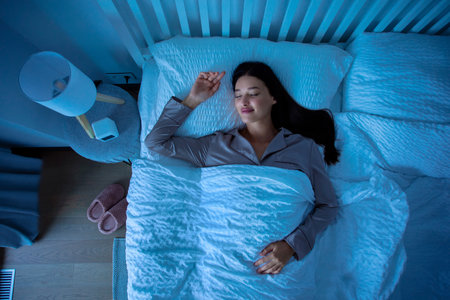 Top view of young calm woman sleeping cozily on bed on white linen at night with blue light shining through the windowの写真素材