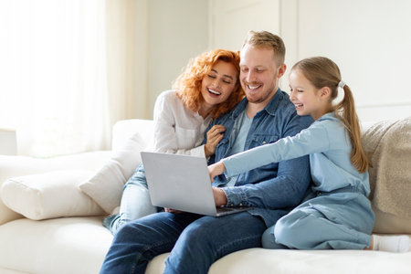 Domestic lifestyle concept. Family of three shopping online via laptop, girl pointing at screen, choosing something with parents, sitting on couchの写真素材