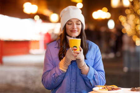 Portrait of European woman enjoying drinking takeaway cup of mulled wine outdoors on street with festive illumination on Christmas Eveの写真素材