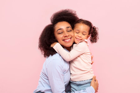 Happy African American mother embracing with adorable little daughter, showing loving feelings, enjoying tender moment, pink studio backgroundの写真素材