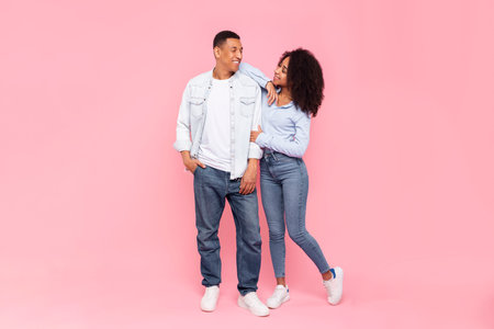 Happy African American couple posing and looking at each other, standing isolated over pink background, full body length shot. Lady leaning on her boyfriends shoulderの写真素材