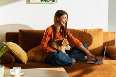 Pretty young lady sitting on sofa with Corgi dog and using laptop, working remotely from home with her lovely petの写真素材