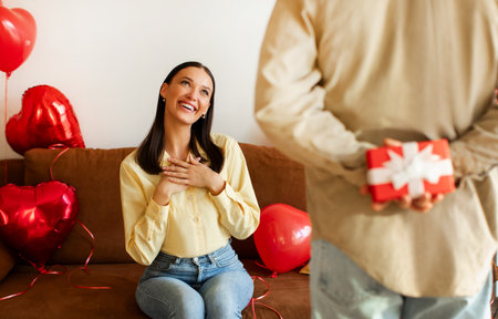 Happy couple celebrating Valentines Day at home, man hiding gift box behind his back, making surprise to wifeの写真素材