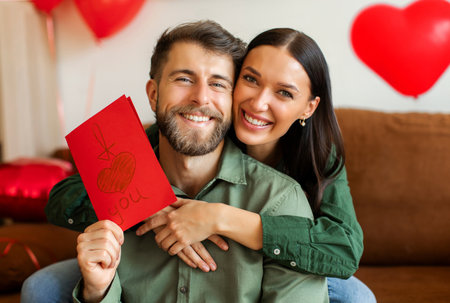 Happy woman giving greeting card to her beloved man with the inscription I love you, smiling together at camera in living room at homeの写真素材