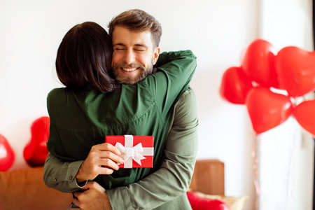 Romantic European couple celebrating Valentines Day, embracing tenderly, man holding gift box, room decorated with heart-shaped balloonsの写真素材